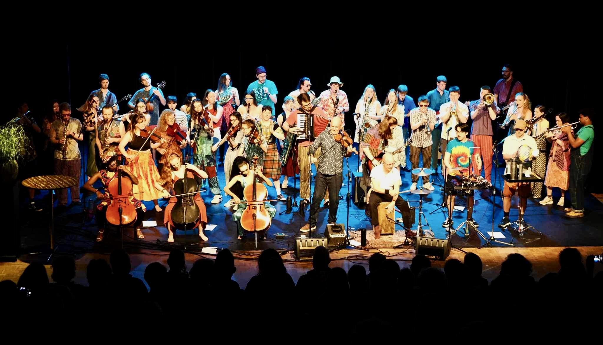 Jonathan Parapadakis playing flute with folk ensemble. Other instruments visible are woodwinds (flute, clarinet, saxophone) and brass (trumpet, trombone, horn) and strings and percussion